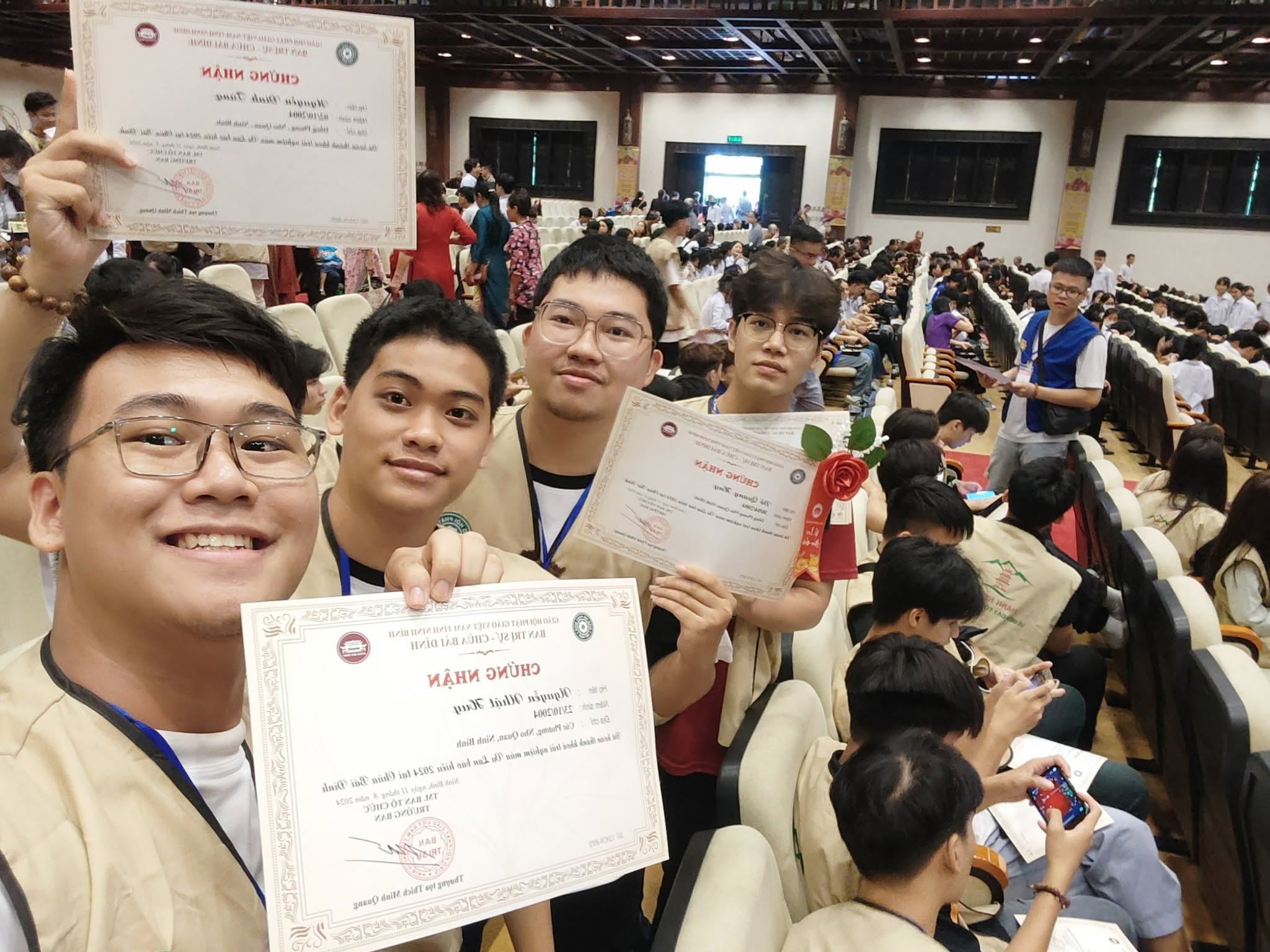 Ảnh selfie bốn bạn mặc áo khoá tu trong hội trường chùa, tay cầm giấy chứng nhận hoàn thành khoá tu mùa hè.