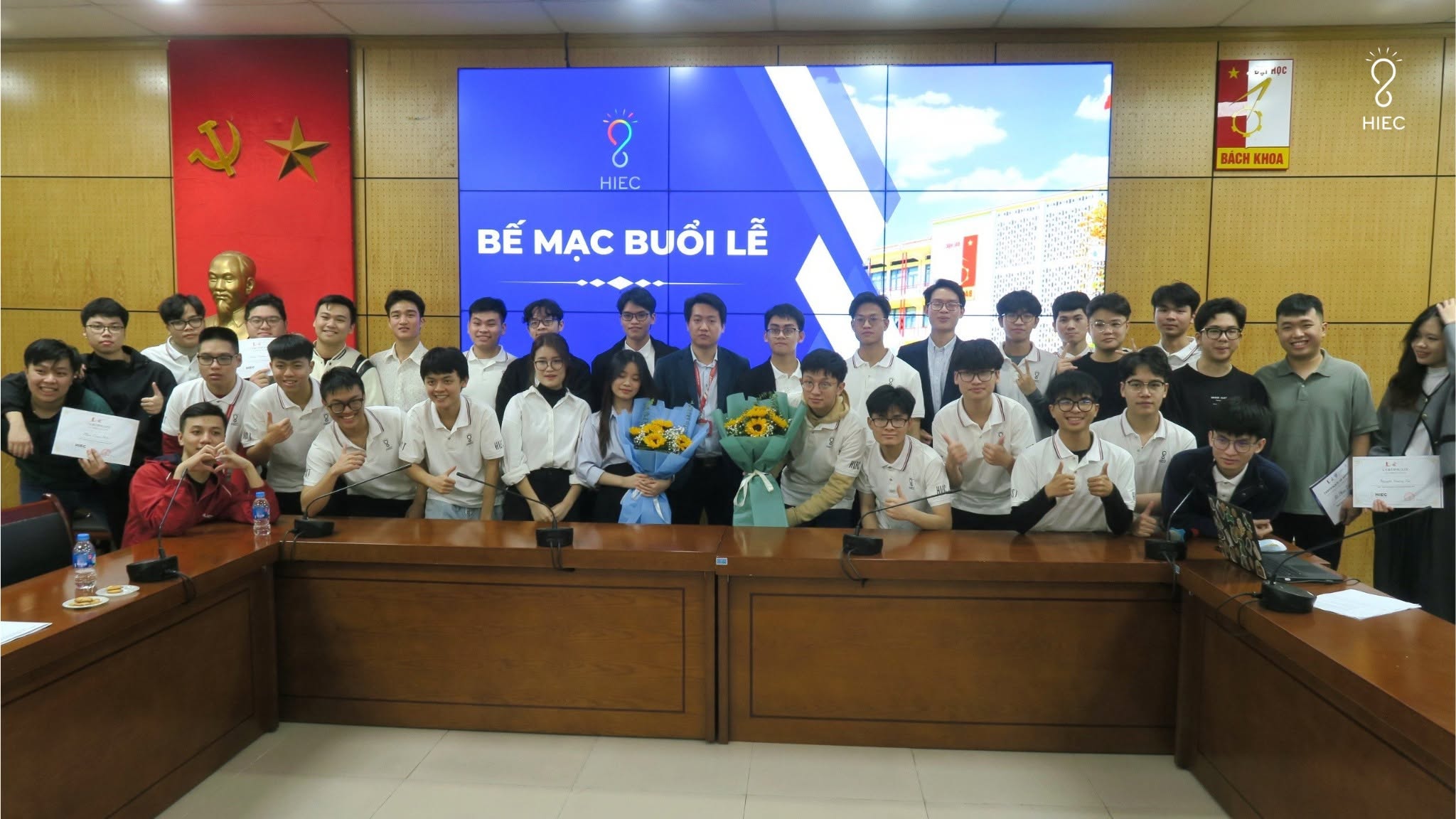 Buổi bế mạc của câu lạc bộ, cả nhóm mặc áo trắng đồng phục đứng chụp chung sau dãy bàn họp, hai bạn ở giữa ôm hoa hướng dương.
