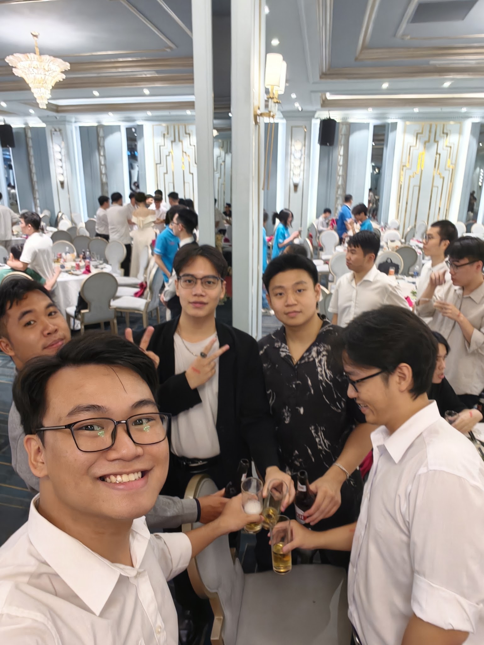 Ảnh selfie cụng ly cùng bạn bè trong sảnh tiệc sau lễ tốt nghiệp, đèn chùm và gương lớn phía sau.