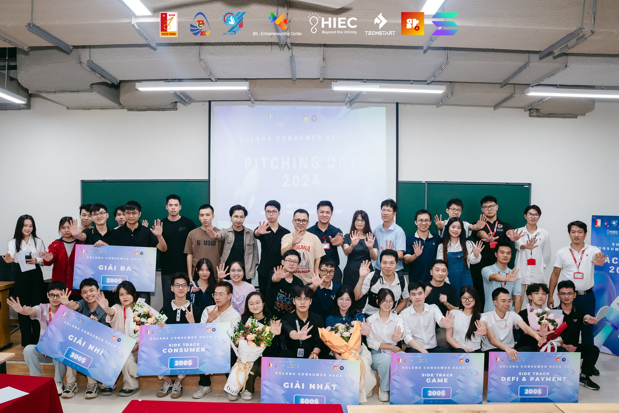 Ảnh tập thể ngày pitching của một hackathon blockchain trong giảng đường, mọi người giơ tay chào máy ảnh.
