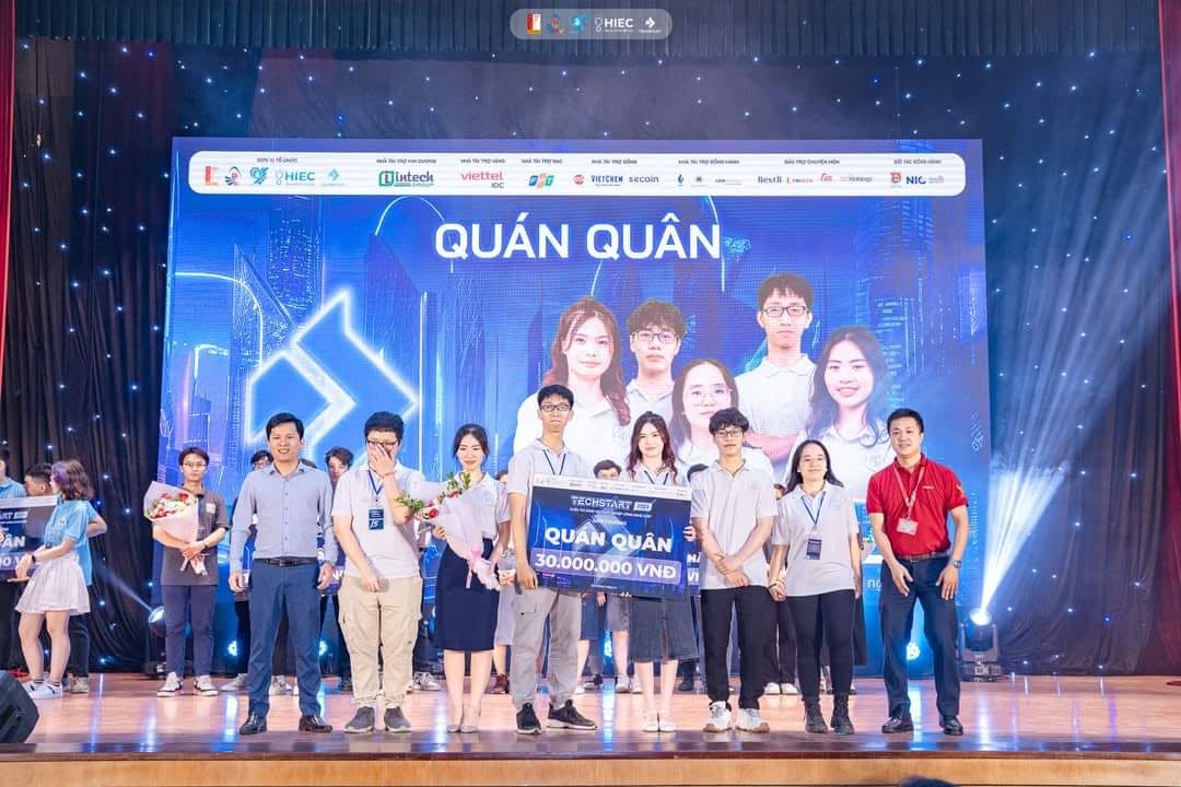 Cả đội đứng trên sân khấu chung kết cuộc thi khởi nghiệp, cầm hoa và bảng tên đội, phông nền xanh thẫm phía sau.