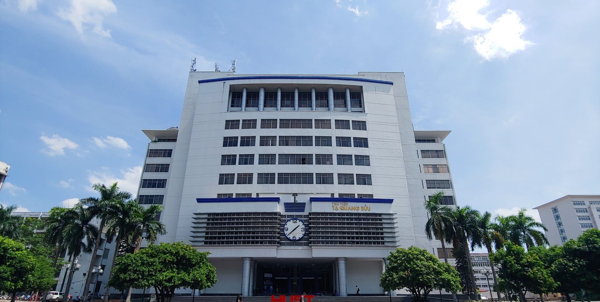 Mặt tiền Thư viện Tạ Quang Bửu nhìn chính diện, hàng cọ hai bên và chữ HUST màu đỏ trước cửa.